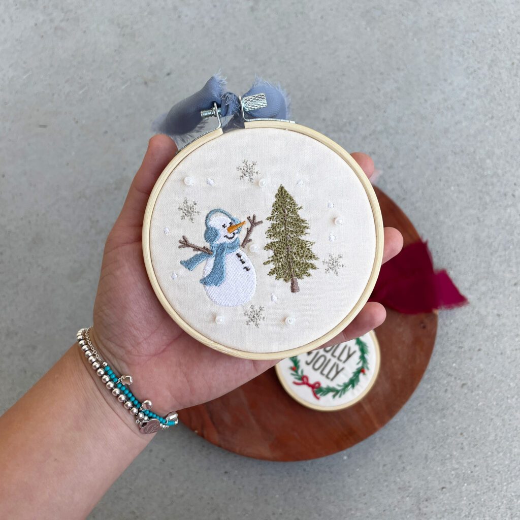 Snowman machine embroidery design displayed as an ornament on a wooden hoop, featuring detailed stitching and festive colors for a charming holiday look.